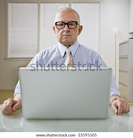 An elderly businessman is working on a laptop in an office.  He is looking at the camera.  Square framed shot.