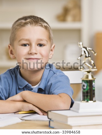 A young boy is seated at a desk with a stack of books and a trophy.  He is smiling at the camera.  Vertically framed shot.