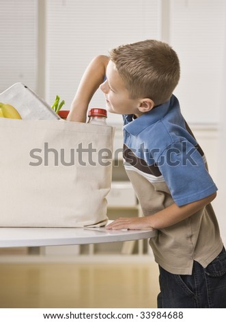 A young boy is getting into the grocery sack on the kitchen counter.  He is looking away from the camera.  Vertically framed shot.