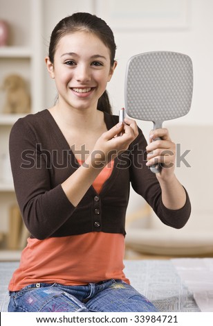 A young girl is looking into a handheld mirror and putting makeup on.  She is smiling at the camera.  Vertically framed shot.