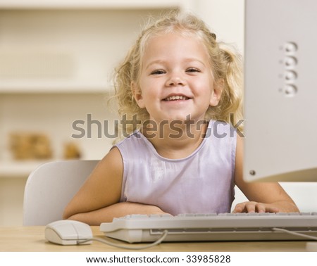 A young girl is seated at a computer desk and smiling at the camera.  Horizontally framed shot.