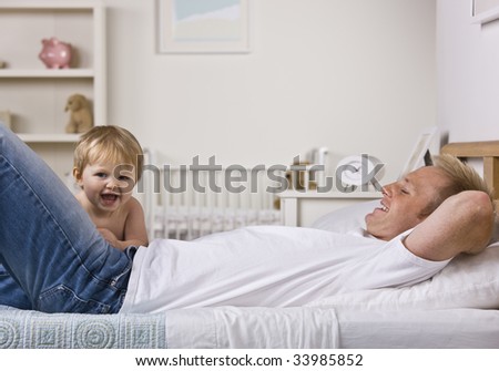 A young father is laying down on a bed and playing with his baby daughter.  Horizontally framed shot.