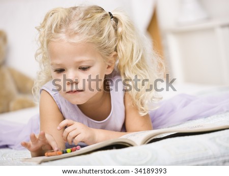 A young girl is laying on a bed and coloring in books.  She is looking away from the camera.  Horizontally framed shot.