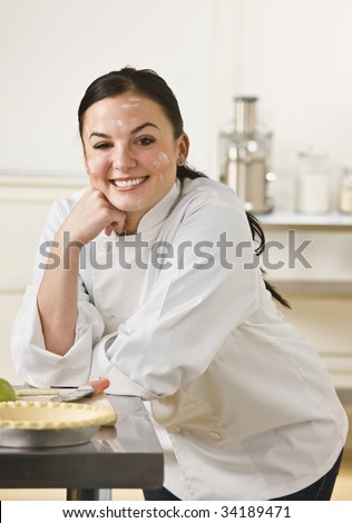 A woman is in a kitchen and baking a pie.  She is smiling at the camera and her face is covered in flour.  Vertically framed shot.