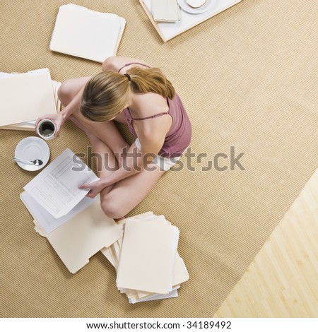 A woman is seated on the floor, looking through files, and drinking coffee.  Square framed shot.