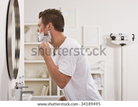 A young man is shaving his face in front of the bathroom mirror.  He is looking away from the camera.  Horizontally framed shot.