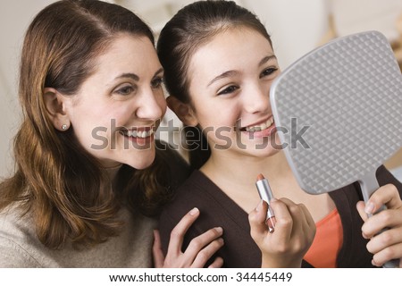 A beautiful young girl holding a mirror and a tube of lipstick with her mother.  They are smiling.  Horizontally framed shot.