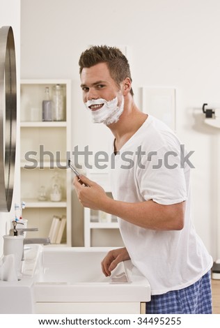 A man is shaving his face in front of the bathroom mirror.  He is smiling at the camera.  Vertically framed shot.