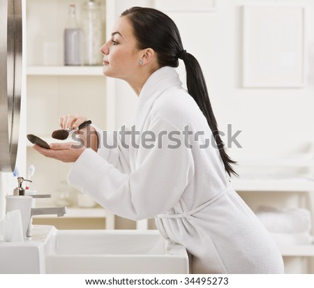A young woman is standing in front of the bathroom mirror and putting on makeup.  Horizontally framed shot.