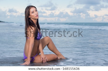 Model at an ocean beach in a purple swimsuit.