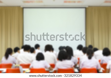 Blurred abstract background of university student sitting in a lecture room in front of the class with white projector slide screen: Blurry view from back of the classroom