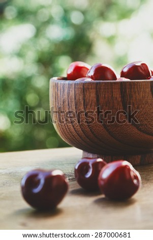 Vertical photo of cherry berries in the wooden bowl on the wooden table on the green bokeh background with three berries out of the bowl