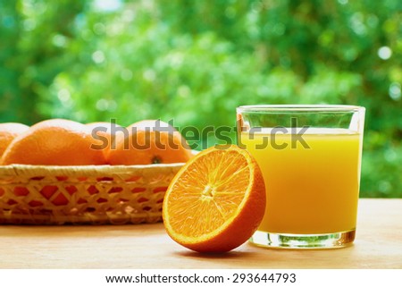 Glass of orange juice, half of orange and basket with oranges on the wooden table on the green blurred background
