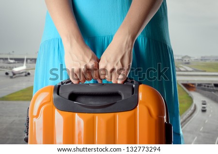 Woman in blue dress holds orange suitcase in hands on airport background.