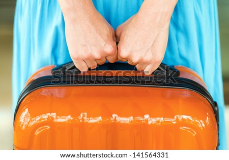 Woman in blue dress holds orange suitcase in hands.