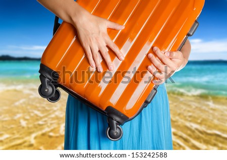 Woman in blue dress holds orange suitcase in hands on the beach background.
