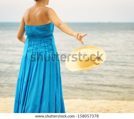 Woman in blue dress throws hat on the beach.
