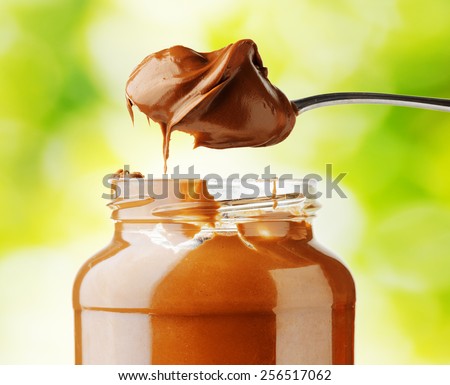 A jar of tasty hazelnut chocolate spread and a little appetizing chocolate paste in a spoon on nature background. Organic healthy food rich in minerals and vitamins. Eco food for sweet breakfast.