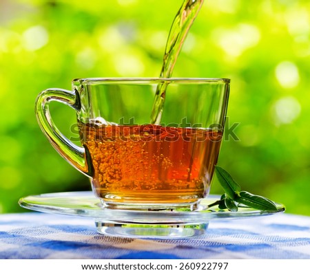 Invigorating fresh aromatic tea pouring into glass cup on the blue and white tablecloth in garden and on nature background.