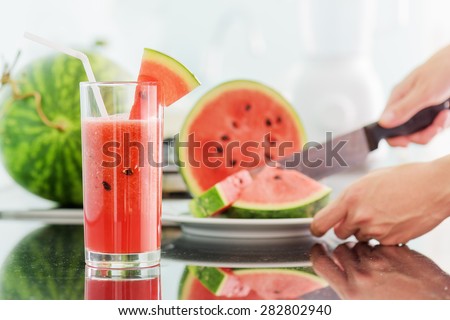 Glass of fresh watermelon juice on table in kitchen. Young woman slicing a ripe watermelon in background. Healthy eco food rich in vitamins. Popular product of organic farming.