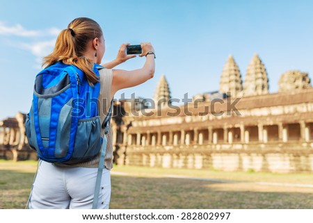 Young female tourist with blue backpack and smartphone taking picture of the eastern facade of the ancient mysterious temple complex Angkor Wat on blue sky background. Siem Reap, Cambodia.