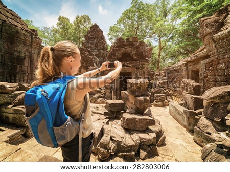 Young female tourist with blue backpack and  smartphone taking picture of old pavilion among mysterious ruins of ancient Preah Khan temple in Angkor. Siem Reap, Cambodia. Toned image.