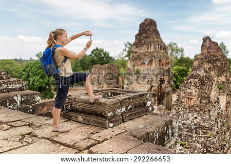 Young female tourist with blue backpack taking picture with smartphone of mysterious ruins from the top of ancient Pre Rup temple Khmer in Angkor on blue sky background. Siem Reap, Cambodia.