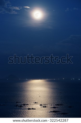 Full moon reflected in water of Nha Trang Bay of South China Sea in Vietnam at night. Marine farms illuminated by beautiful mysterious moonlight. Island and lights of ships are visible in background.