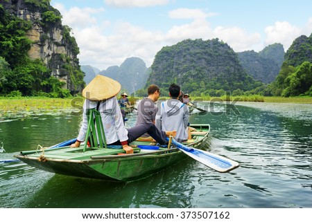 Tourists traveling in boat along the Ngo Dong River at the Tam Coc portion, Ninh Binh Province, Vietnam. Rower using her feet to propel oars. Scenic landscape formed by karst towers and rice fields.