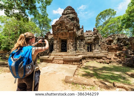 Young female tourist with smartphone taking picture of the gopura under blue sky near the entrance to ancient Preah Khan temple in Angkor. Siem Reap, Cambodia.