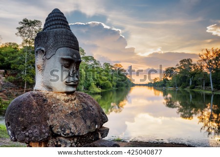 Stone Asura on causeway near South Gate of Angkor Thom in Siem Reap, Cambodia. Beautiful sunset over ancient moat in background. Angkor Thom is a popular tourist attraction.