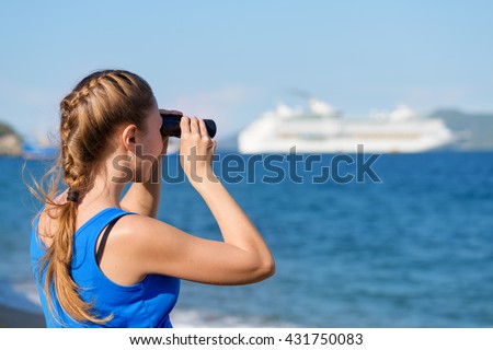 Young female tourist looking through binoculars at white cruise ship (liner) and enjoying beautiful sea view. Woman wearing blue dress. Her hair braided in French plait. Outdoor portrait in summer.