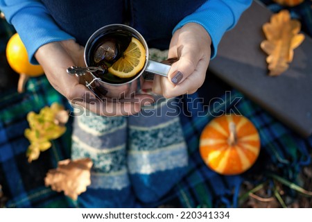Female hands with hot tea cup and female legs in warm knitted socks on plaid background, book, autumn leaves and small pumpkin, top view