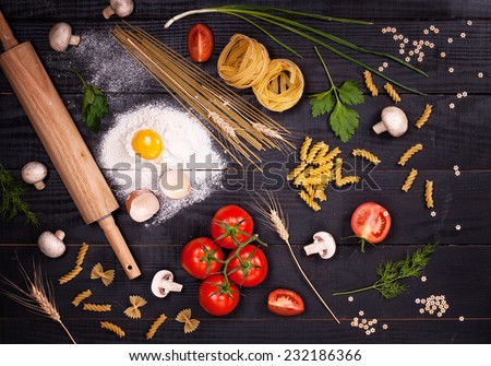 Raw pasta, tomatoes,mushrooms, flour and eggs on black wooden table background, top view