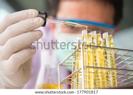 Laboratory pipette with drop of yellow liquid over glass test tubes for an experiment in a science research lab .Man wears protective goggles