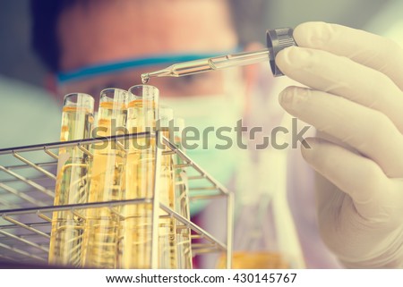Laboratory pipette with drop of yellow liquid over glass test tubes for an experiment in a science research lab .Man wears protective goggles
