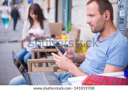 Woman with cell phone and the man with laptop and mobile  phone sitting in a cafe.  Secluded alley in a background.