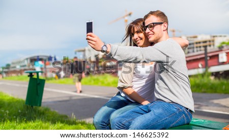 Two Smiling young people taking a picture with mobile phone. They are sitting on a bench.
