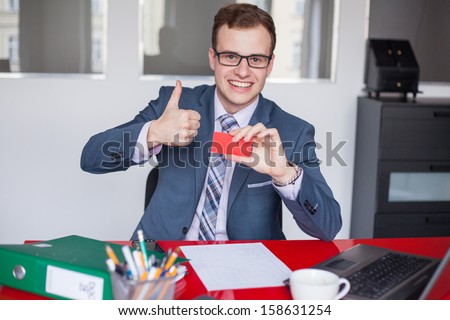 Young businessman holding mobile phone and red business card. He is sitting in his office.