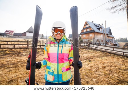 Beautiful girl with a full ski suit, ski goggles and ski helmet in front of mountain houses. He is holding a skis