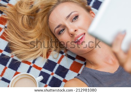 Sensual blonde woman lying in park on blanket. She is using white tablet pc. Outdoor photo. She looks relaxed