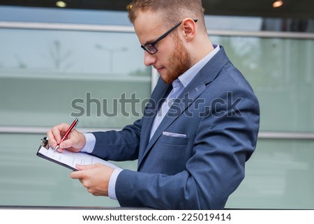 Portrait of young businessman with beard standing in front of office block. He is signing a contract. Outdoor photo