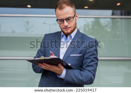 Portrait of young businessman with beard standing in front of office block. He is signing a contract. Outdoor photo