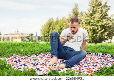 Bearded man sitting in park on blanket. He is using mobile phone. Outdoor photo. Negative emotion.