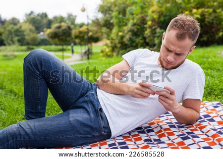 Bearded man sitting in park on blanket. He is using mobile phone. Outdoor photo. Negative emotion.