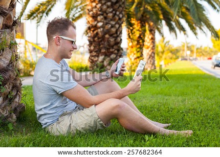 Young tourist sitting under palm tree with mobile phone and tablet pc. Positive emotion. Vacation time.