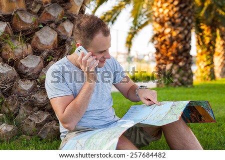 Young tourist sitting under palm tree with mobile phone and tablet. He is looking at the map. Positive emotion. Vacation time.