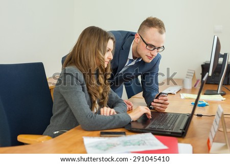 Man and woman spending time in the office. Woman sitting behind the computer, man standing next to her and showing something on the screen.