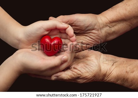 little girl and elderly woman keeping red heart in their palms together, symbol of care and love