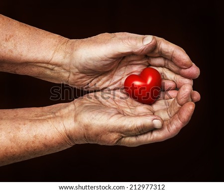 elderly woman keeping red heart in her palms isolated on dark background, symbol of care and love